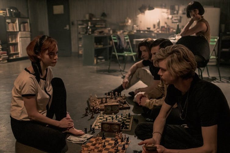 A young woman in front of a line of young men playing chess.