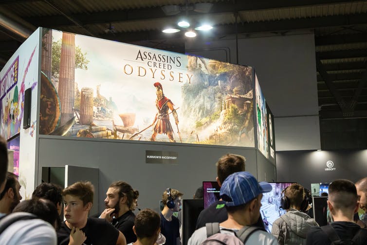 A giant billboard showing a gladiator from the video game Assassin's Creed overlooks a crowd of people.