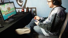 A young man sits in front of a computer with a game console.