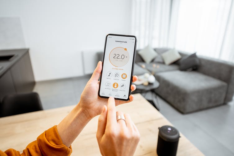 A hand holds a smartphone in a living room. A temperature control app is open on the screen.