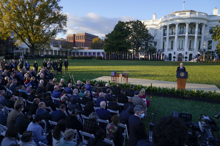 With the White House in the distance, President Biden speaks at a lectern before a crowd of people