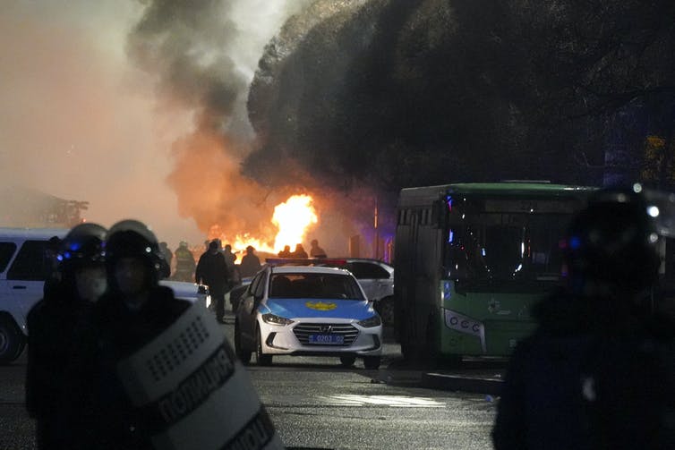 A city street with police in riot gear, a police car, a bus and flames in the background