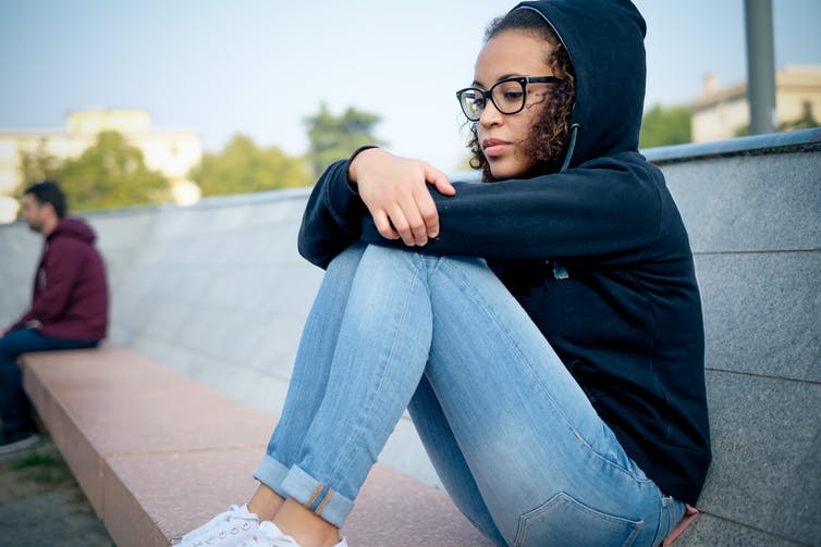 A young woman wearing glasses, jeans and a hoodie sits alone on a bench with her arms resting on her knees