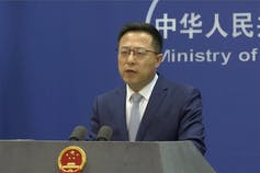 a man in a dark business suit speaks at a podium with a red and gold emblem on the front