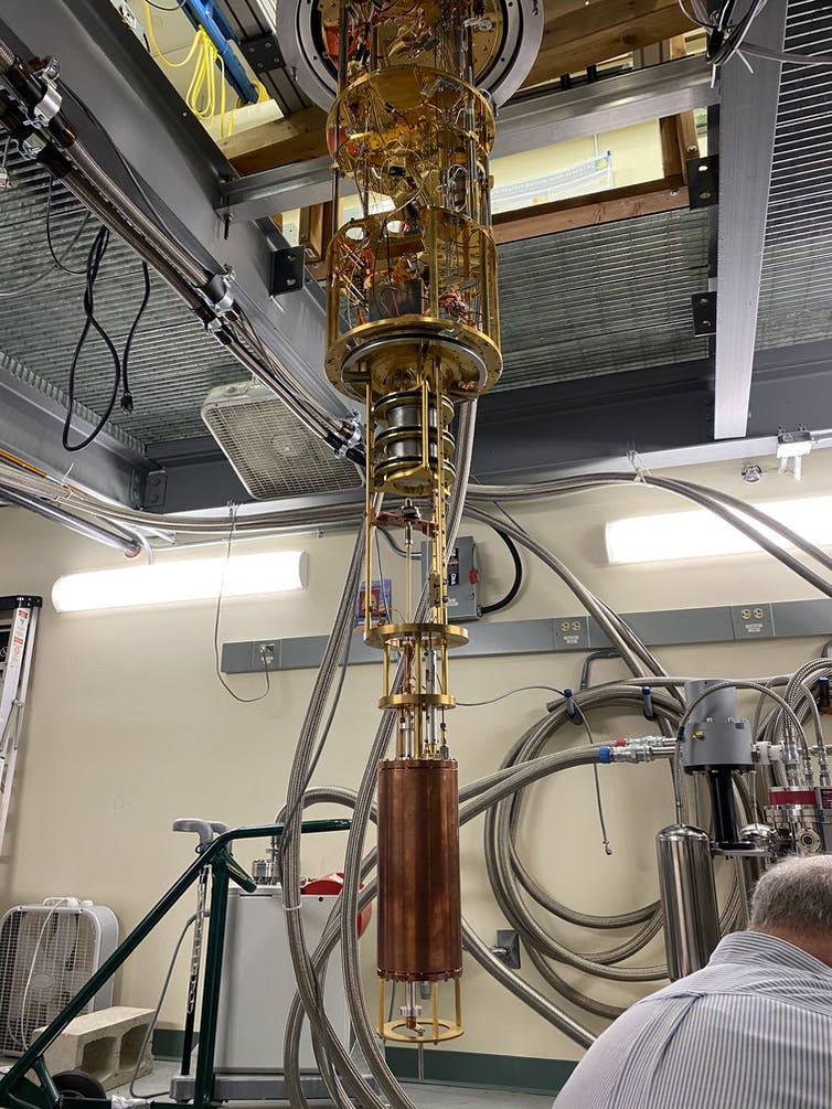 The HAYSTAC detector, a large copper cylinder connected to a gold-plated assembly of tubes and wires hanging from the ceiling of a lab.