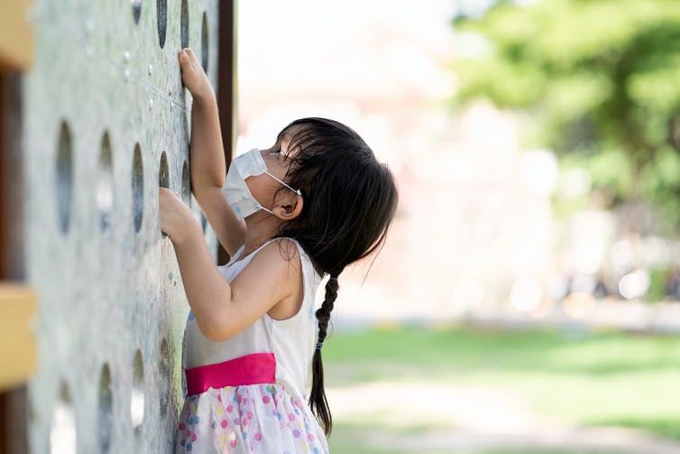 A child wearing a face mask looks up at a climbing wall.