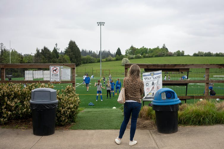 A woman in a leoplard print jacket seen by herself near a covid sign watching a soccer game.