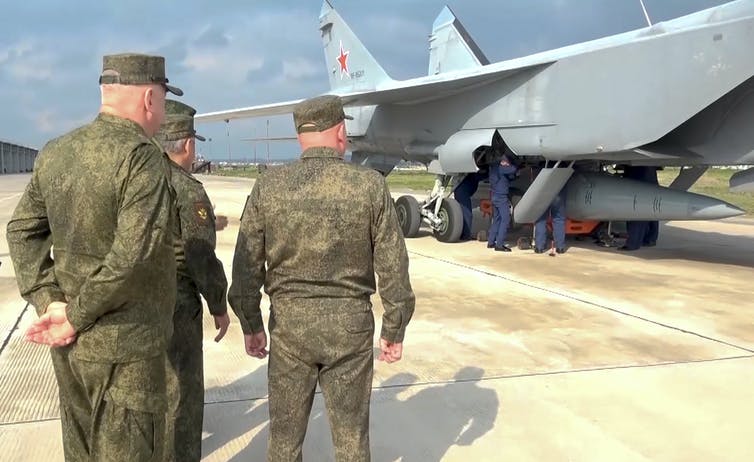 men in military uniforms watch technicians work on a missile beneath a military jet plane on a tarmac