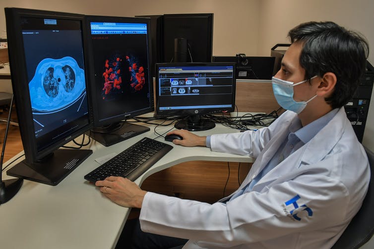 A man in a white lab coat and wearing a facemask looks at several computer screens showing medical images