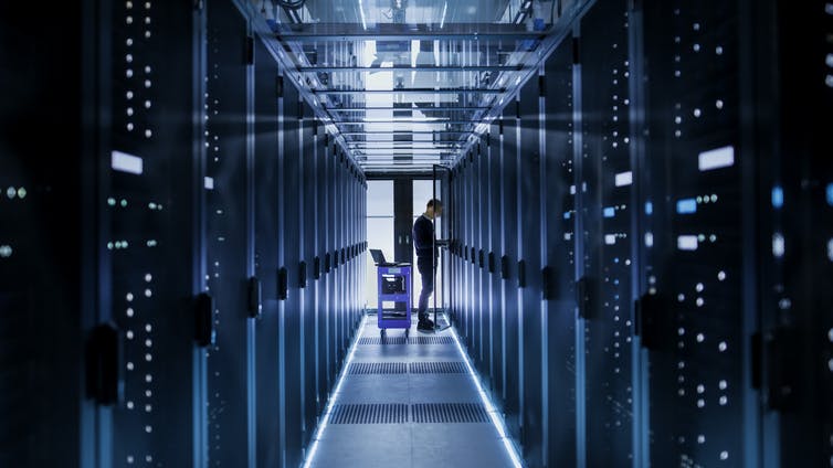 A man stands at the end of long, bluish room with walls of tech on either side.