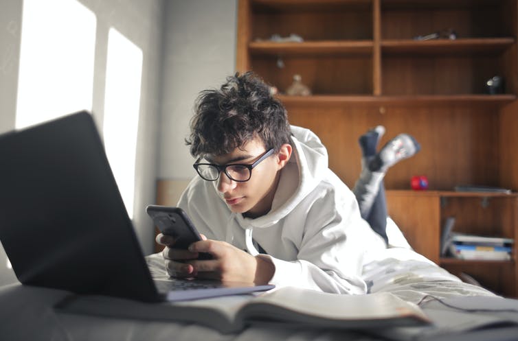 A teenage boy lies on his bed using his smartphone, with a laptop open.