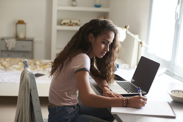 A girl sits at a desk in front of her laptop and writes on a notepad