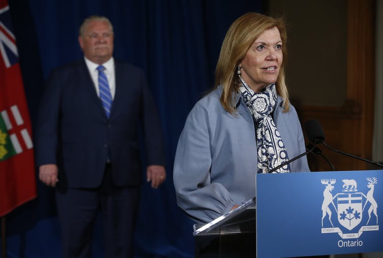 Ontario Premier Doug Ford looks on as Ontario Health Minister Christine Elliott stands at a podium