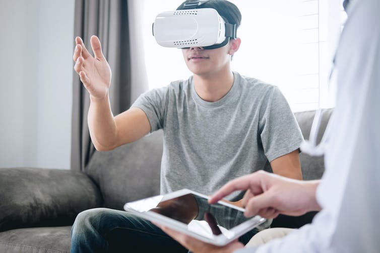 A man wearing a VR headset sits next to someone holding a tablet.