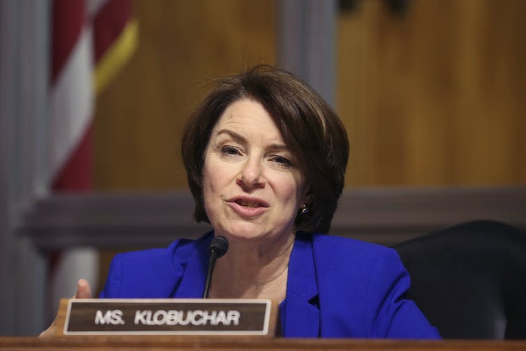 A middle-aged white woman with brown hair wearing a blue blazer sits behind a nameplate