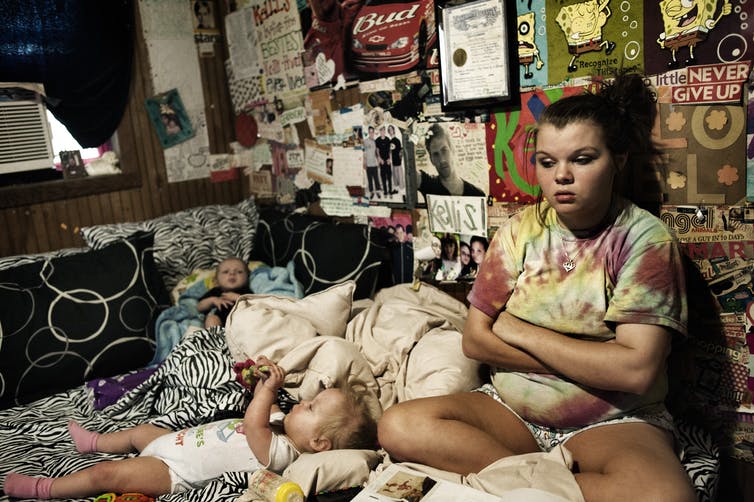 A teenage girl sits in a bedroom with posters on the wall, with her arms crossed. Two young children lie on the bed around her.