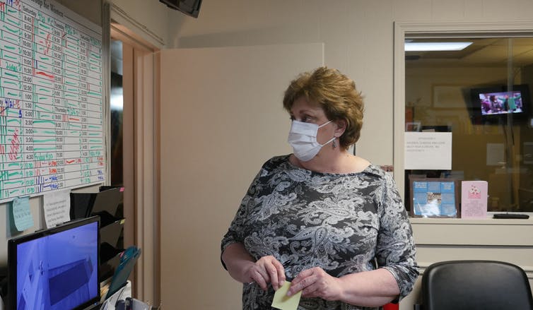 A brown haired woman wearing a gray patterned shirt and a mask looks at a whiteboard full of information in an office.