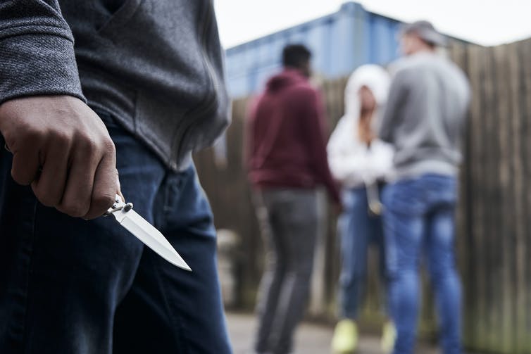 Close up of a young man's hand holding a knife, while a group of three other young people are huddledin the distance