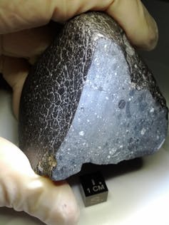 A photograph of a gloved hand holding a pyramid-shaped rock, blackish grey on the outside, sliced open to reveal white crystals inside.