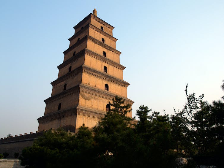 A tall Chinese pagoda against a blue sky rises above a row of trees in the foreground