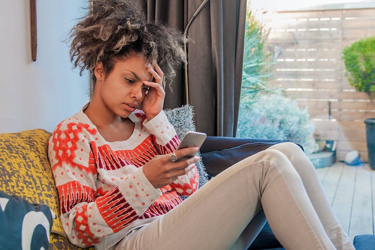 A woman in casual clothes on a sofa, looking at her phone with her hand to her face, looking concerned