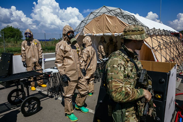 A soldier stands in the foreground as a half dozen people in hazmat suits and gas masks stand near stretchers outside a large tent