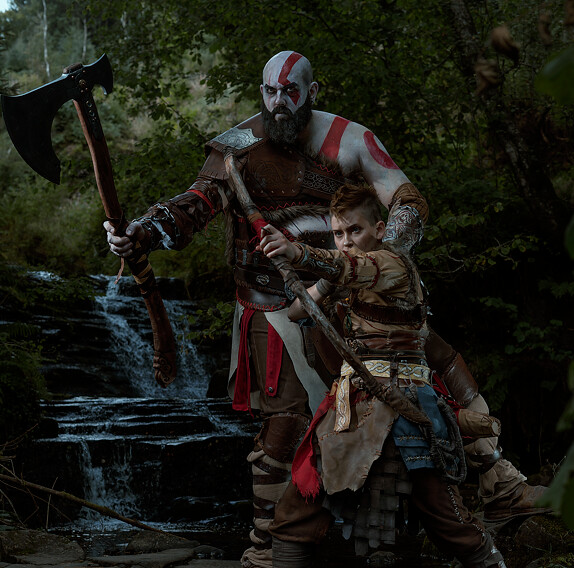 This image shows cosplayers portraying Kratos and Atreus in front of a stream. Kratos has his axe raised looking off at an unseen target while Atreus points his bow towards it, ready to fire.