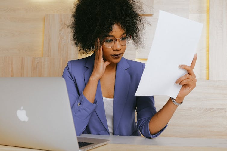 Woman in business suit at laptop reading sheet of paper