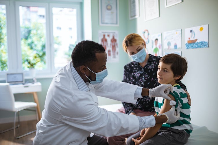 A young boy sits on an exam table with his mother soothing him while a doctor puts a Band Aid on his arm after giving him a shot.