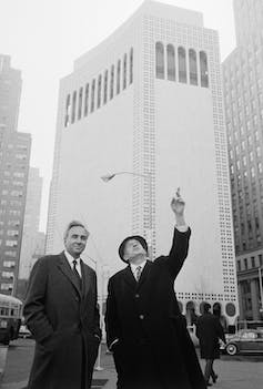 Two white men in suits, ties and coats stand next to each other in New York in front of a tall building. One of them is pointing and looking up