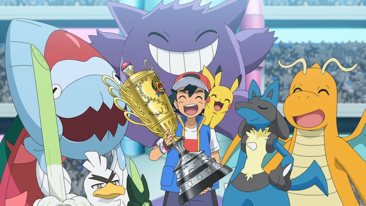 Ash Ketchum stands holding a big cup trophy, with Pikachu on his shoulder and other large Pokemon surrounding them