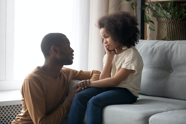 A man sits in front of his daughter, who is sitting on a couch, holding her hand and talking to her