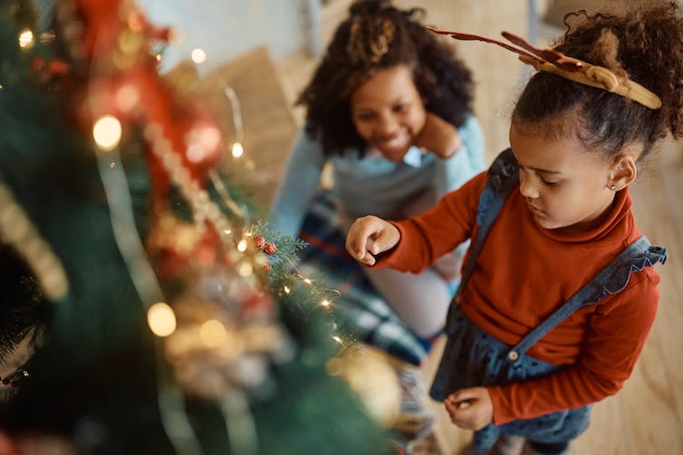 A mother watches her daughter hand an ornament on a Christmas tree