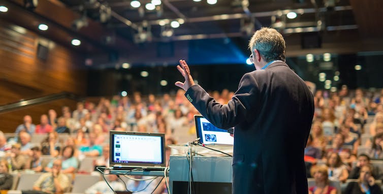 a man gives a lecture while reading from two laptop screens