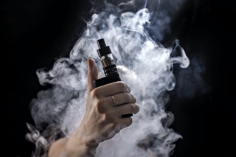 A hand holding a vaping device surrounded by smoke against a black background