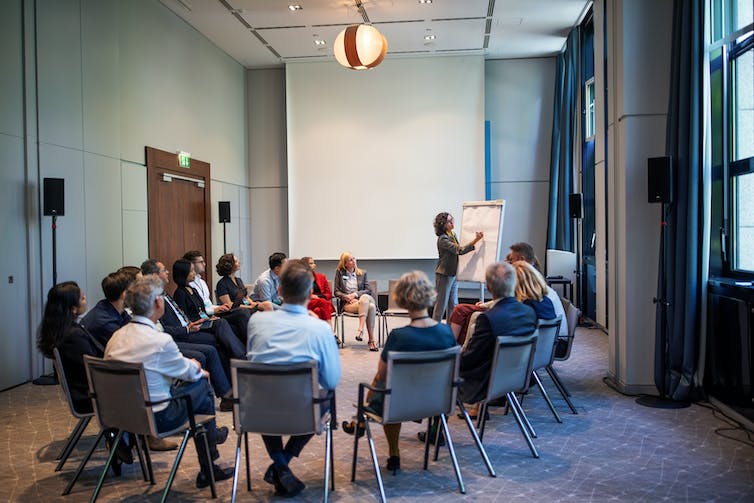 A group of people sitting in a semi-circle watching a woman give a presentation.