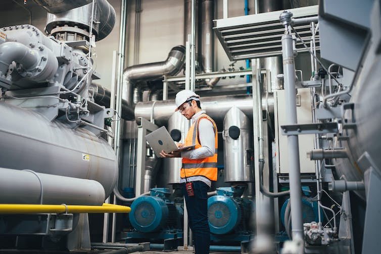 Workman holding a laptop next to some boiler pipes