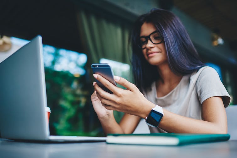 A woman wearing glasses sits at a laptop while looking at a smartphone and wearing a smart watch.