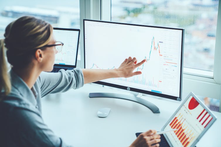 Woman looking and pointing at financial charts, screen and laptop, window in the background.