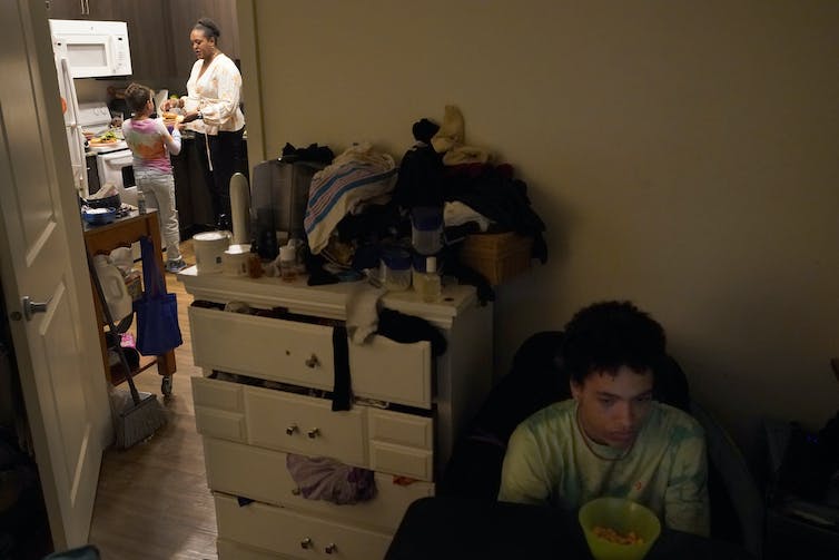 A child seen in front of a glowing computer screen while a parent is in a kitchen with another child.