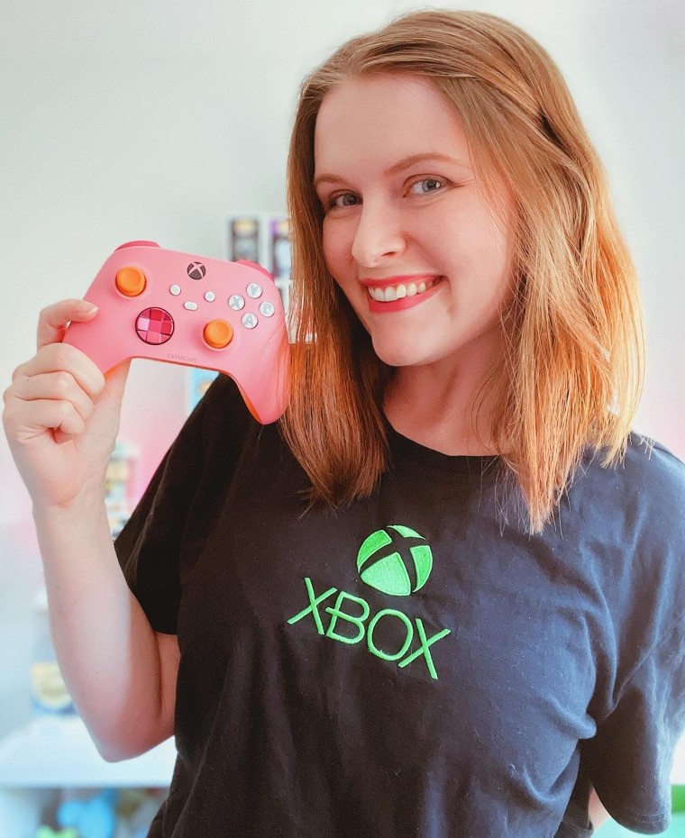 ctrlaltcraft smiles in an Xbox t-shirt, holding up a pink and orange Xbox controller next to her face.