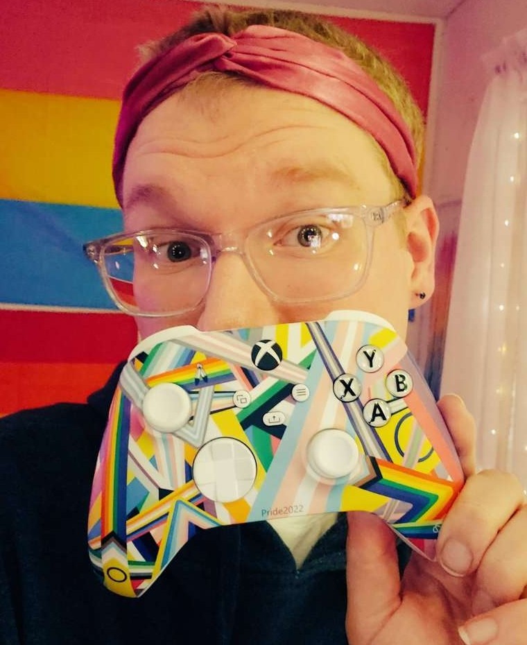 ItsMileyGirl holds up an Xbox Pride controller in front of their mouth, with a Pride flag in the background. She is wearing transparent glasses and a pink headband.