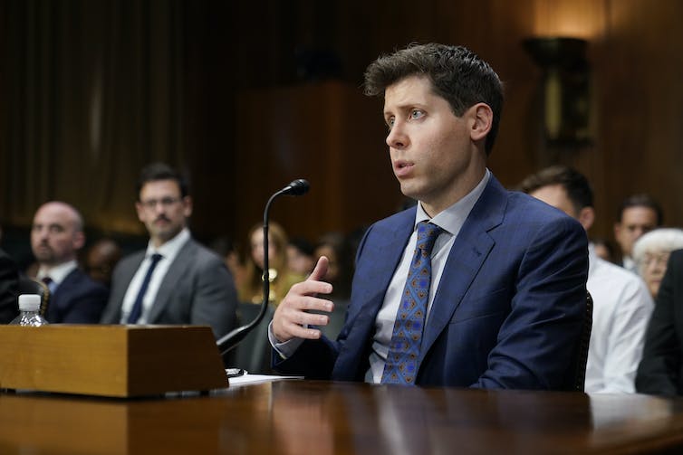 Man in a blue suit is seated at a desk speaking into a microphone with people seated behind him.