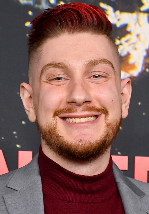 Shawn stands in front of a step and repeat, smiling at the camera and wearing a grey suit over a maroon shirt.