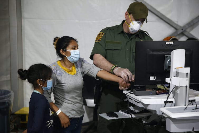 A woman wears a face mask and stands next to a computer next to a small child. A man in a green uniform and a mask holds her finger down near the computer.