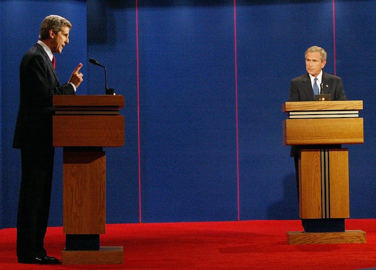 Two men in dark suits debating each other from different lecterns.