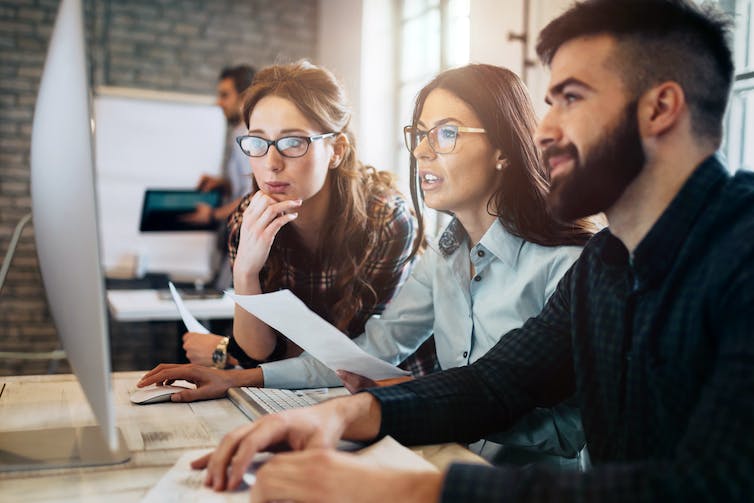 Three colleagues, two women and one man in a cool office sit side-by-side and work on a computer together