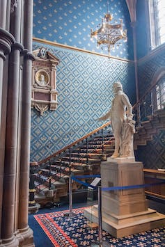 White statue surrounded by a staircase with blue and red carpet, blue and gold wallpapered walls, gold chandelier..