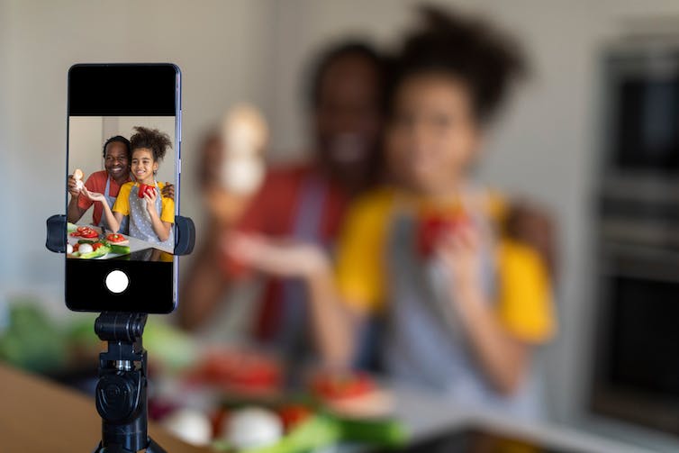 A man and a young girl preparing food in a kitchen while a smartphone films