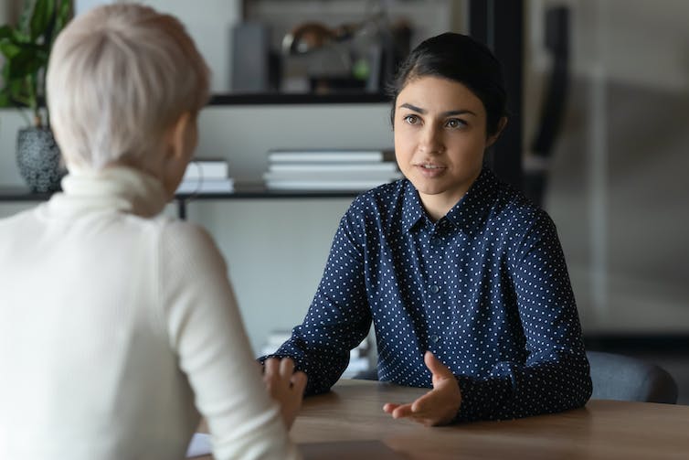 Two women have a conversation at a desk. One woman has her back to the camera.